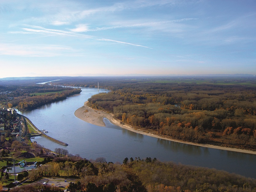 Aerial view of the River Danube surrounded by forest. On one river bank there is a large sediment deposit and on the other one can see a few houses of the town of Hainburg, Austria.